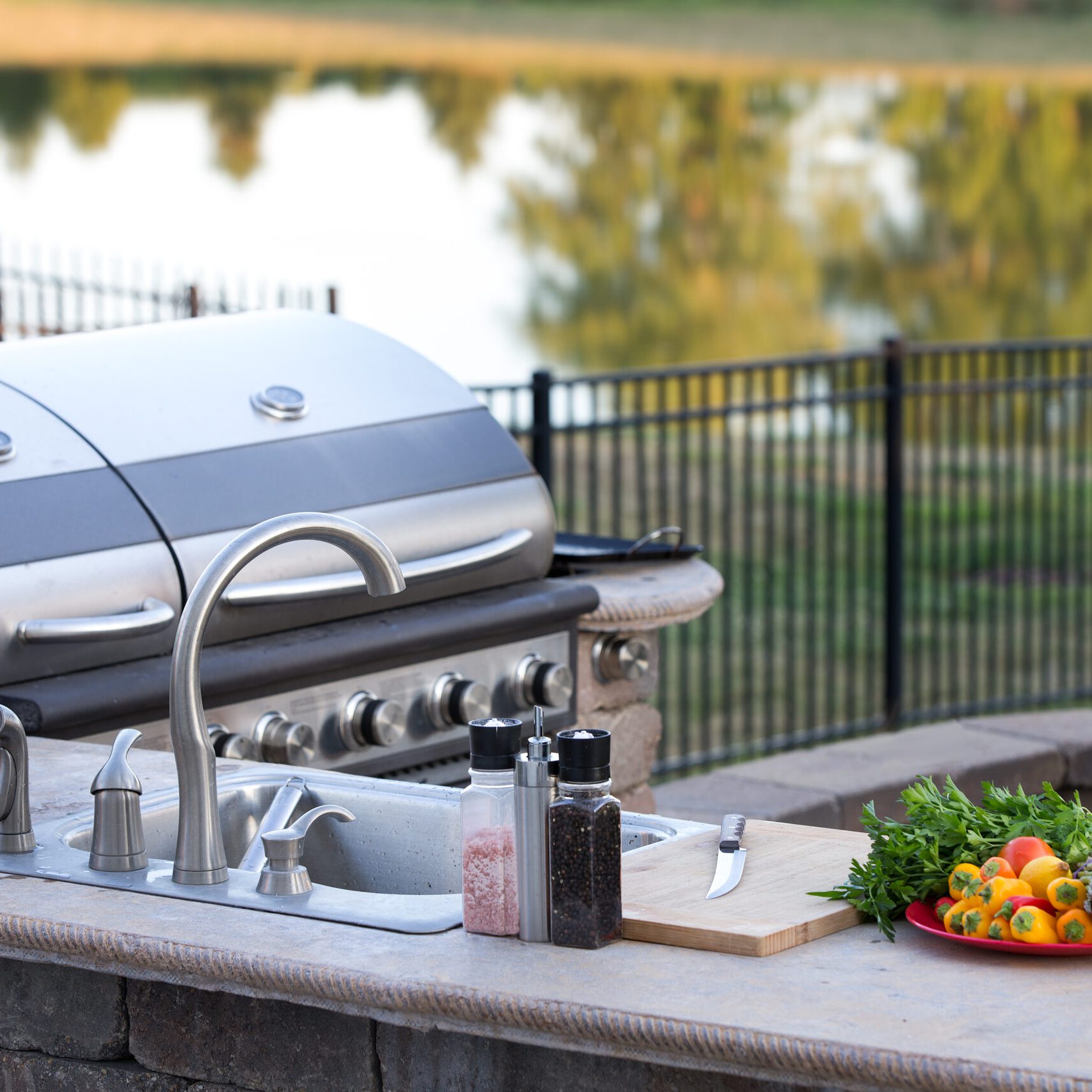 Preparing a healthy summer meal in an outdoor kitchen with gas barbecue and sink on a brick patio overlooking a tranquil lake with tree reflections